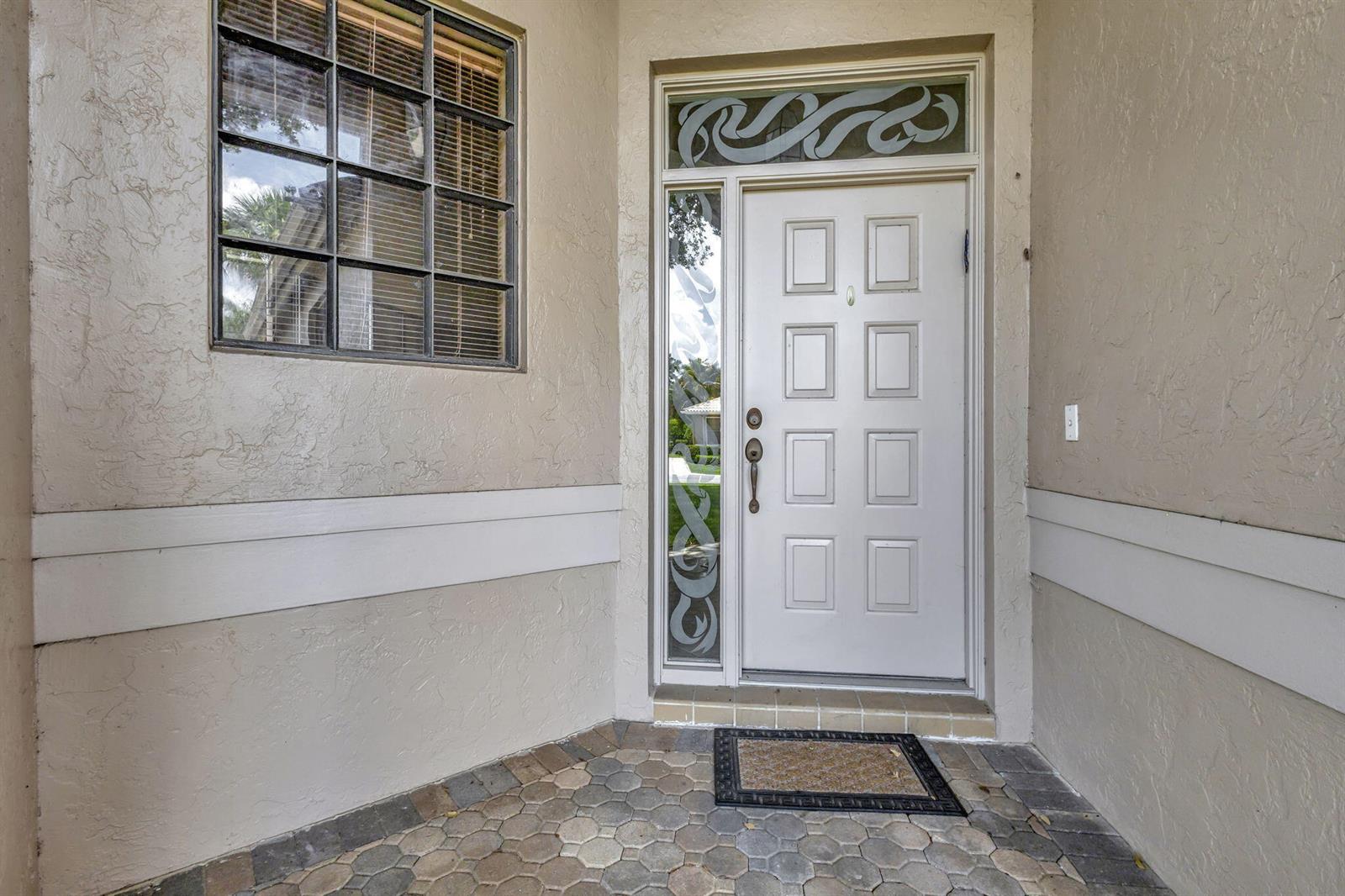 A white front door with a sidelight and transom window sits on a paved porch — 4528 Barclay Fair Way, Lake Worth FL 33449, LAKE WORTH, FL