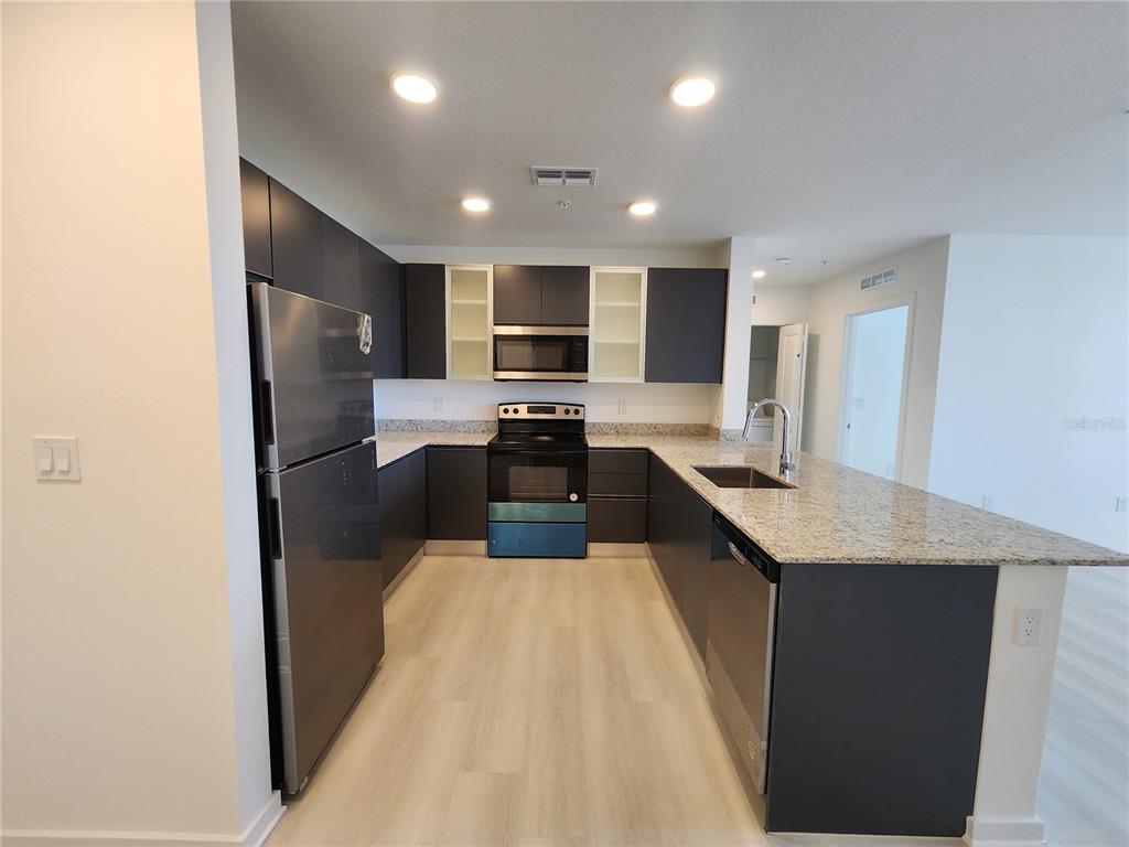 A modern kitchen features dark cabinetry, granite countertops, stainless steel appliances, and light wood flooring — 5579 MILLENIA PARK BLVD UNIT 509, ORLANDO, FL