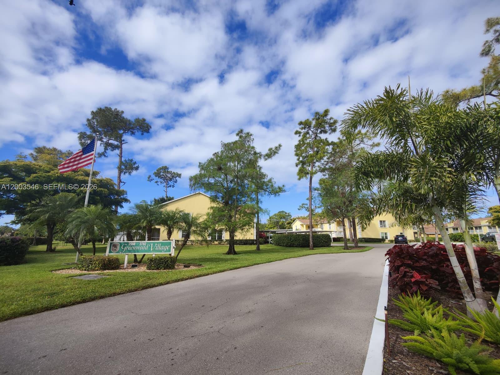 A paved driveway leads toward yellow apartment buildings at the Greenwood Village community entrance — 2668 Kings Lake # 2668, Naples FL 34112, NAPLES, FL