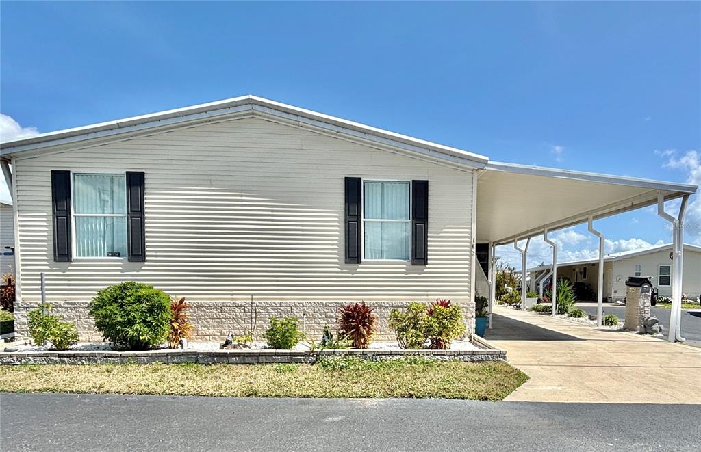 A light-colored mobile home features black shutters, a stone-textured base, and an attached carport — 136 AMERICANA COURT NE UNIT 230, ST PETERSBURG, FL