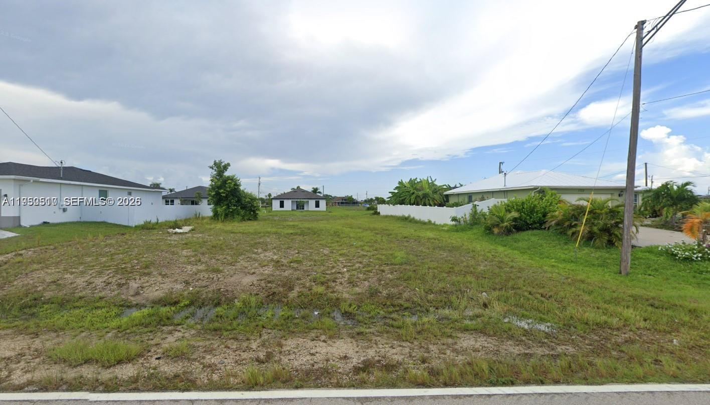 A vacant, grassy residential lot is situated between two existing houses under a cloudy sky — 1718 N Nelson Rd, Cape Coral FL 33993, CAPE CORAL, FL