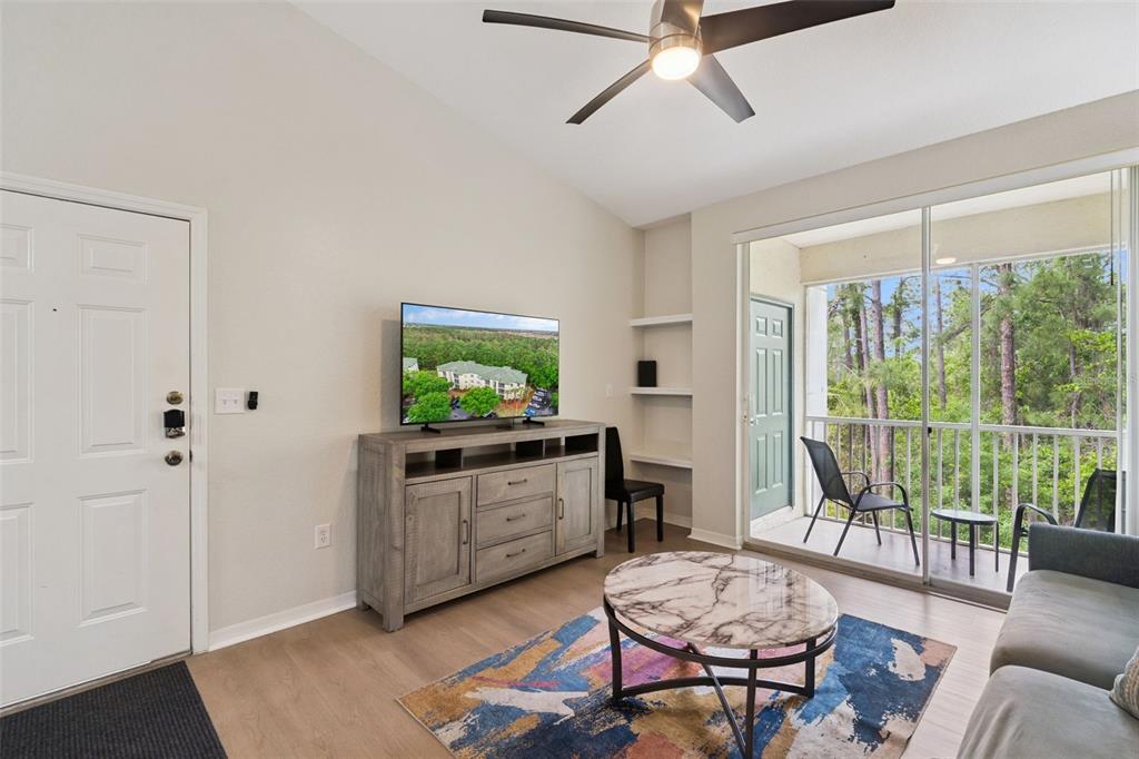 A living room features light wood floors, a ceiling fan, a television, and a balcony — 8921 LEGACY COURT UNIT 18-308, KISSIMMEE, FL
