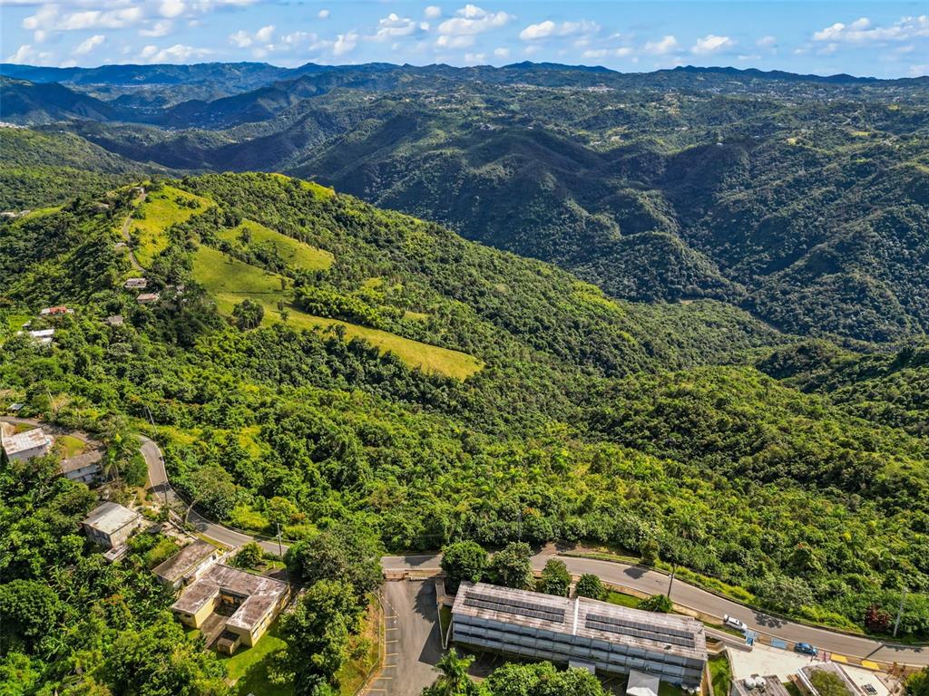 An aerial view shows a multi-story building with solar panels nestled in a mountainous landscape — Carr. 156 KM 20.1 INTERIOR BO. HONDURAS, BARRANQUITAS, PR