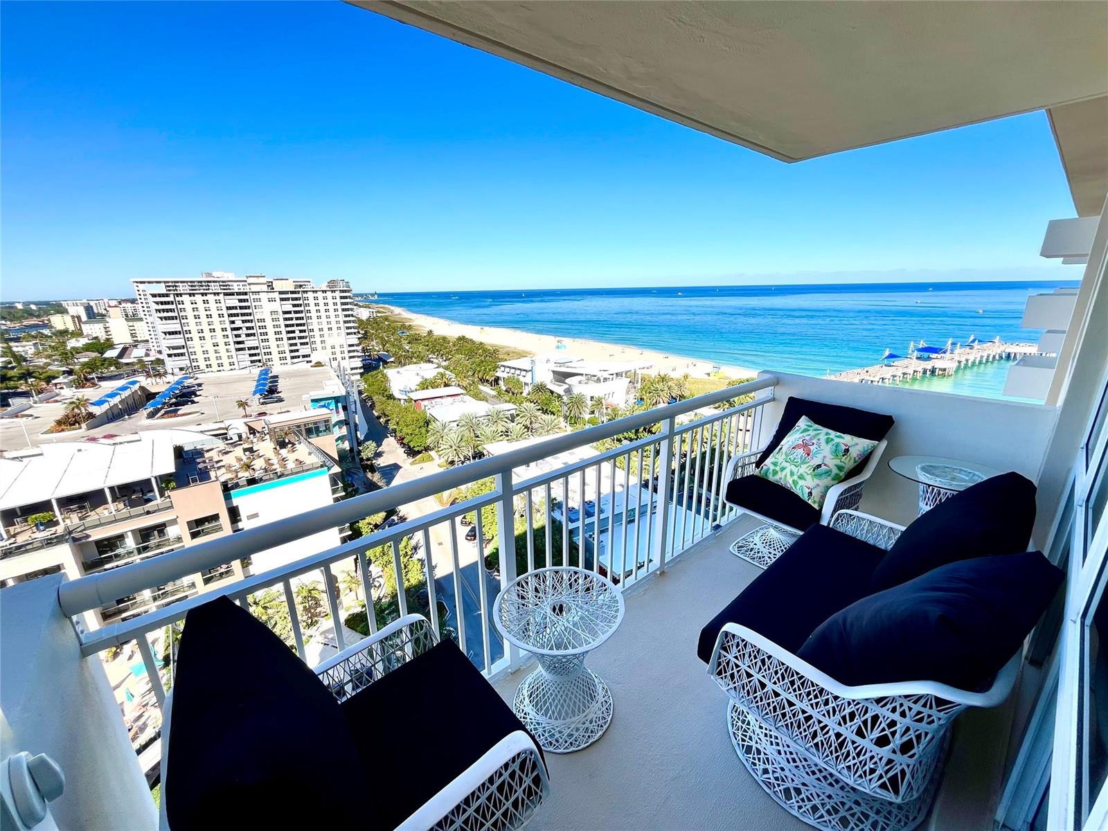 A balcony with white wicker furniture overlooks a beach, ocean, and coastal city buildings — 133 N Pompano Beach Boulevard # 1506, Pompano Beach FL 33062, POMPANO BEACH, FL