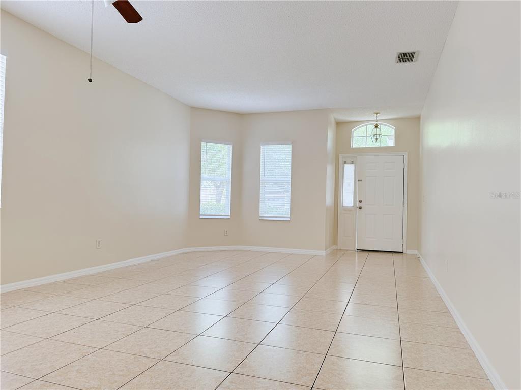 An empty living room features beige tile flooring, light-colored walls, and a front entry door — 4932 BELLTHORN DRIVE, ORLANDO, FL