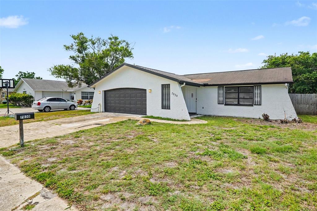 A single-story white house with a dark garage door and a brown shingled roof — 7030 INGLESIDE DRIVE, PORT RICHEY, FL