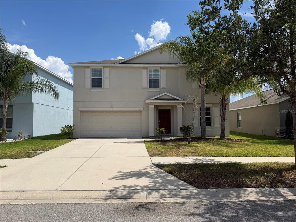 A two-story beige house with a two-car garage and a front porch under a blue sky — 10013 CRESTED FRINGE DRIVE, RIVERVIEW, FL