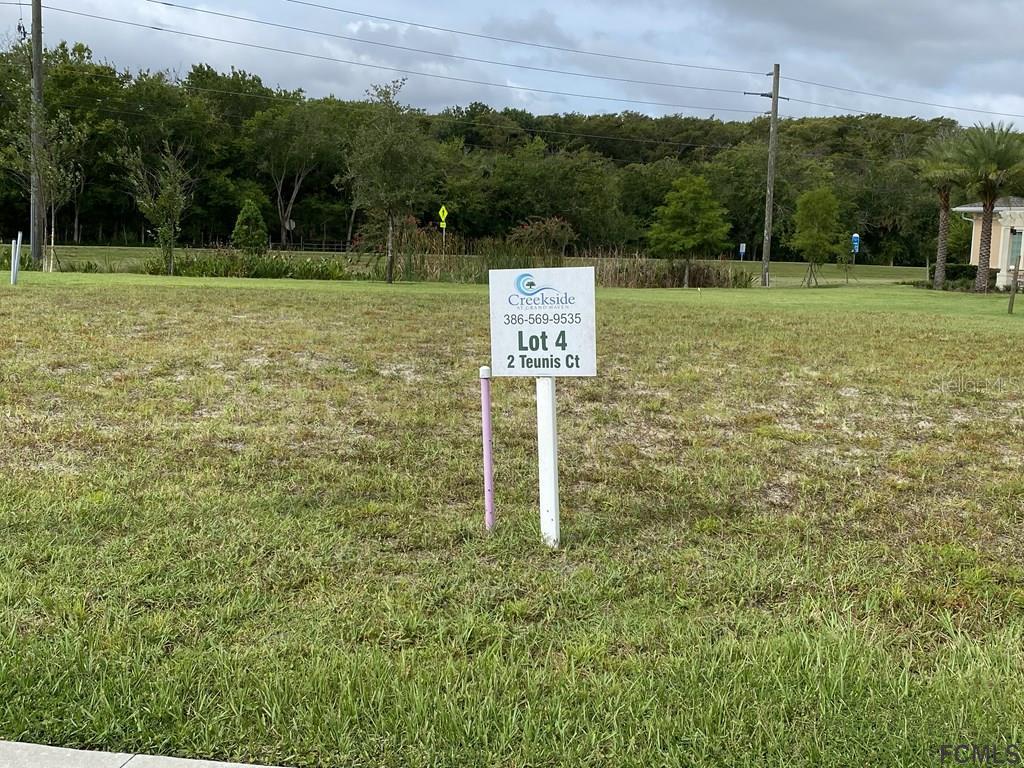 A real estate sign stands on a grassy vacant lot with trees in the background — 2 TEUNIS COURT, PALM COAST, FL