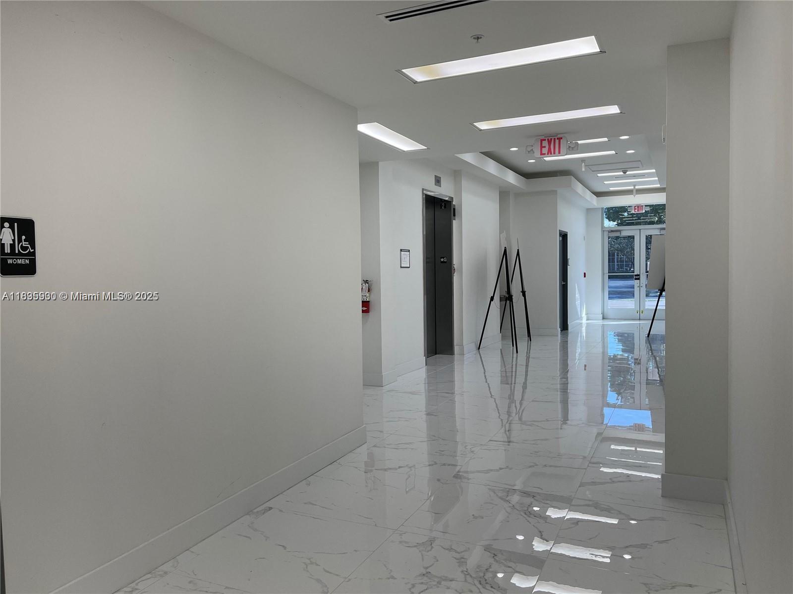 A bright hallway features white walls, marble-patterned tile flooring, an elevator, and glass exit doors — 10450 NW 33rd St # 102, Doral FL 33172, DORAL, FL