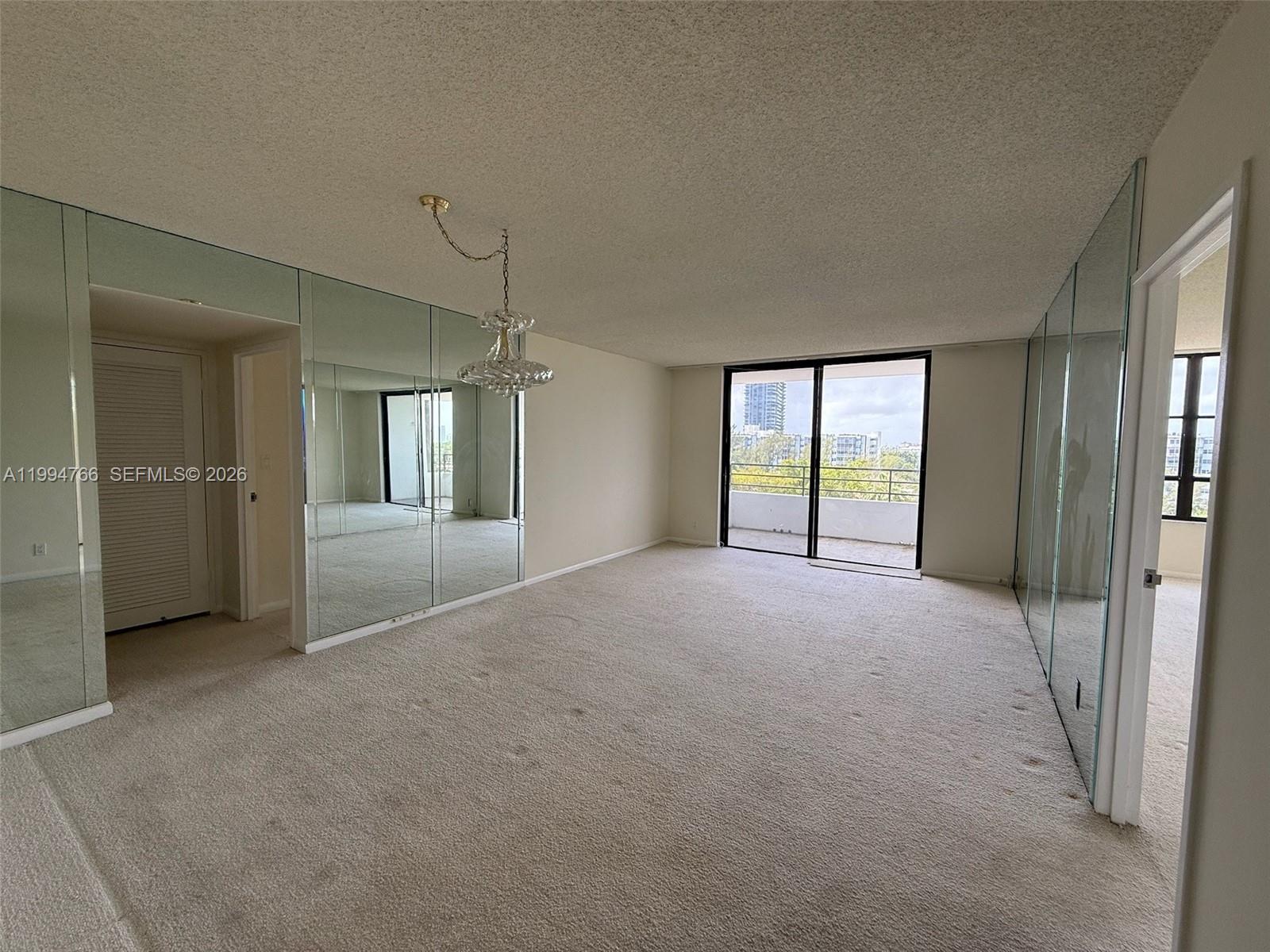 An empty living room features beige carpeting, mirrored walls, a chandelier, and a sliding glass door — 500 Three Islands Blvd # 314, Hallandale Beach FL 33009, HALLANDALE BEACH, FL