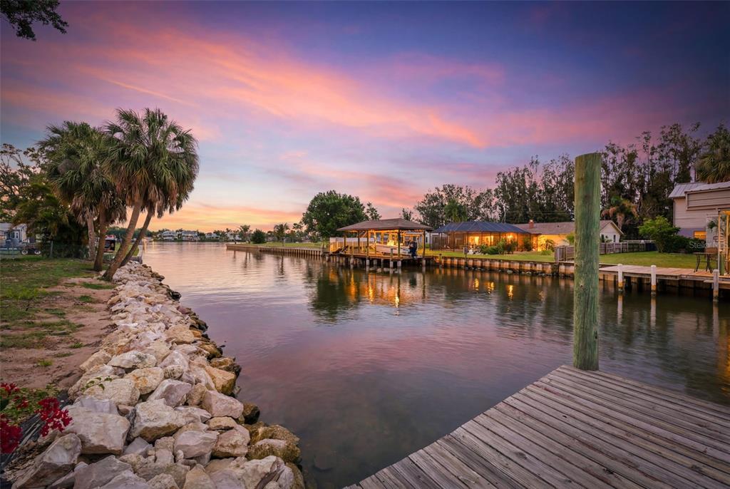 A wooden dock overlooks a canal at sunset with a boat lift and nearby houses — 9910 PENINSULAR DRIVE, GIBSONTON, FL
