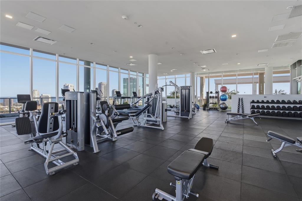A modern gym features various exercise machines, dumbbell racks, and floor-to-ceiling windows with city views — 777 3RD AVENUE N UNIT 1202, ST PETERSBURG, FL