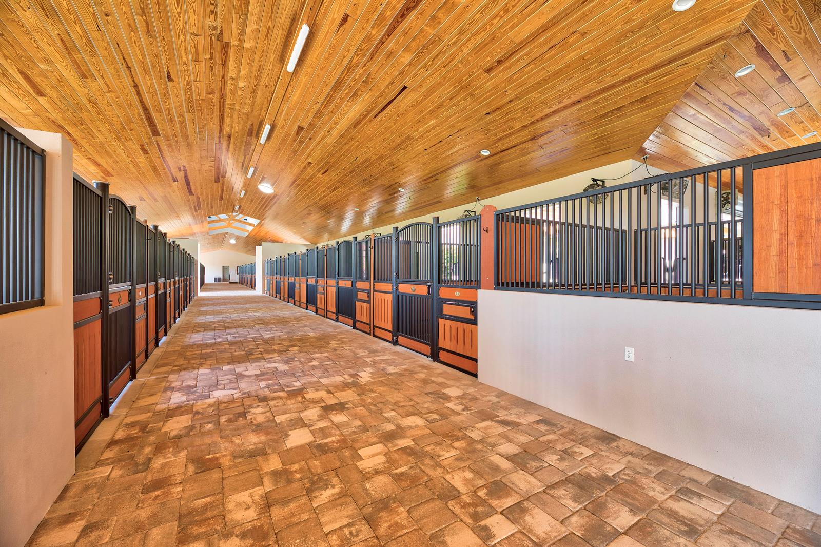 A long horse stable aisle features brick flooring, wood-paneled ceilings, and multiple metal stall doors — 5219 130th Avenue S, Wellington FL 33449, WELLINGTON, FL