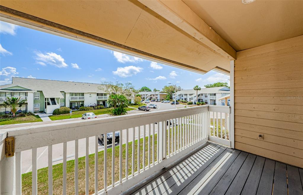 A covered balcony with a wooden floor and white railing overlooks an apartment complex parking lot — 455 ALT 19 S UNIT 214, PALM HARBOR, FL