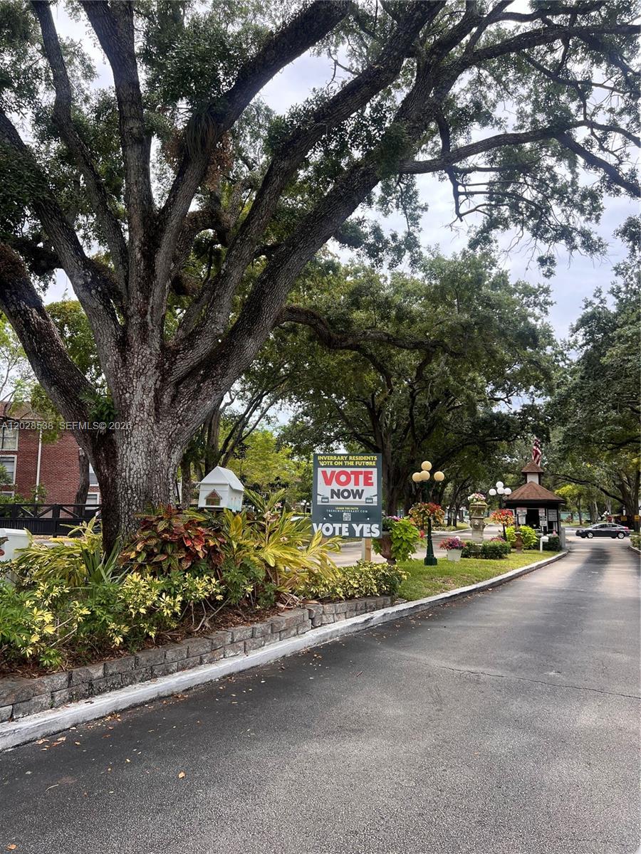 A paved driveway leads toward a gated entrance under large trees with a political sign — 3760 Inverrary Dr # 2A, Lauderhill FL 33319, LAUDERHILL, FL