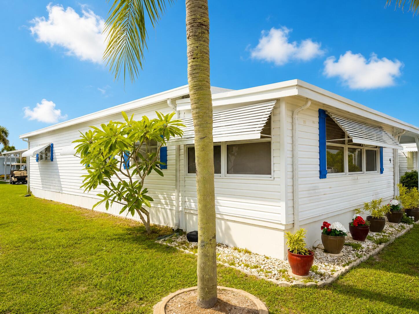 A white mobile home with blue shutters and potted flowers sits on a grassy lot — 3500 S Kanner Highway Lot 31, Stuart FL 34994, STUART, FL