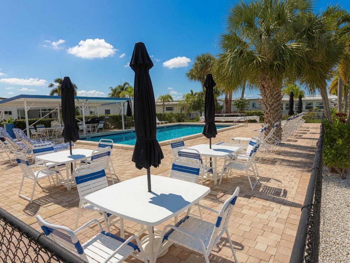A swimming pool area features white tables with black umbrellas and blue-and-white patio chairs — 3500 S Kanner Highway Lot 31, Stuart FL 34994, STUART, FL