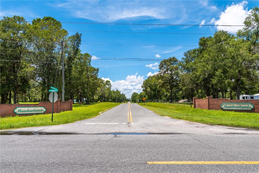 A paved road leads into a residential neighborhood entrance marked by brick walls and signs — Lot 5 SW MEADOWLANDS DRIVE, LAKE CITY, FL