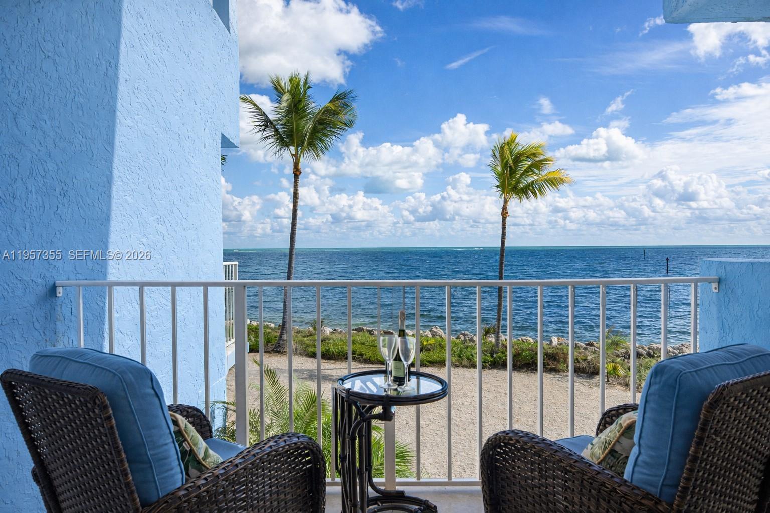 A balcony with two wicker chairs and a small table overlooks the ocean and palms — 1501 Ocean Bay Dr # A1, Key Largo FL 33037, KEY LARGO, FL