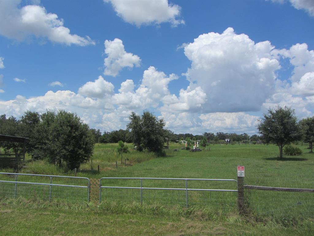 A metal fence borders a grassy field with scattered trees under a cloudy blue sky — 46365 STATE ROAD 64 E, MYAKKA CITY, FL