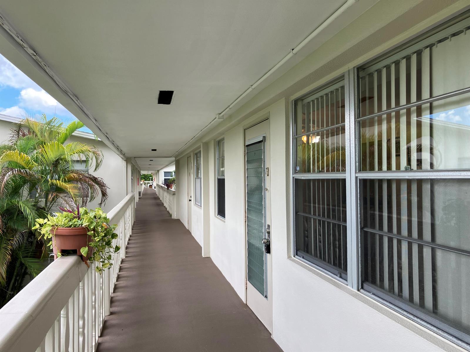 An outdoor walkway leads past apartment doors and windows with a potted plant on the railing — 103 Prescott E, Deerfield Beach FL 33442, DEERFIELD BEACH, FL