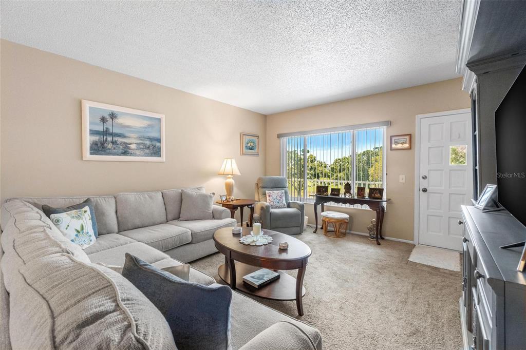 A living room features a sectional sofa, coffee table, armchair, and a view of trees — 2440 WORLD PARKWAY BOULEVARD UNIT 52, CLEARWATER, FL