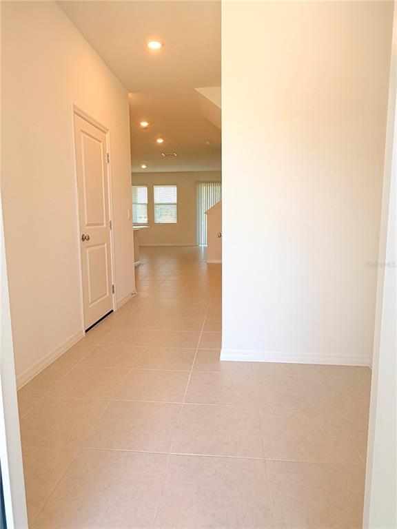 A hallway with light-colored tile flooring leads toward an open-concept living area with windows — 36162 OBSIDION LANE, ZEPHYRHILLS, FL