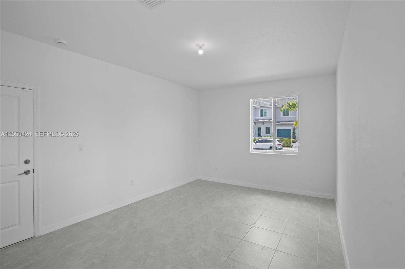 An empty room features white walls, gray tile flooring, and a single window — 715 SE 15th St, Homestead FL 33034, HOMESTEAD, FL