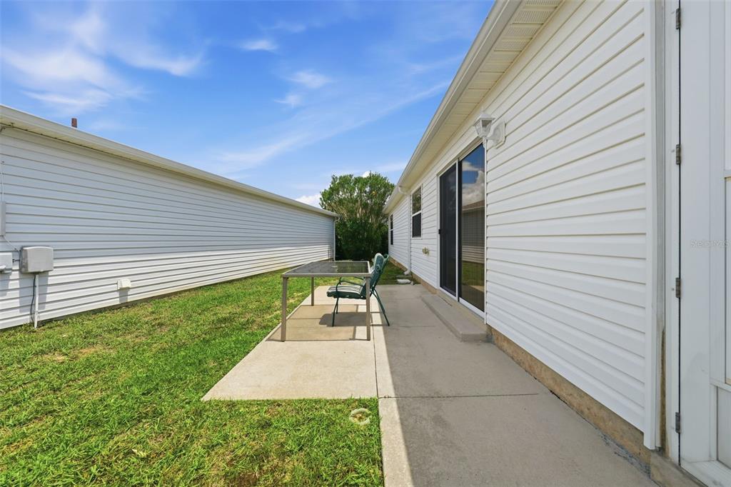 A concrete patio with a table and chairs sits between two white vinyl-sided houses — 1614 GIBBES WAY, THE VILLAGES, FL