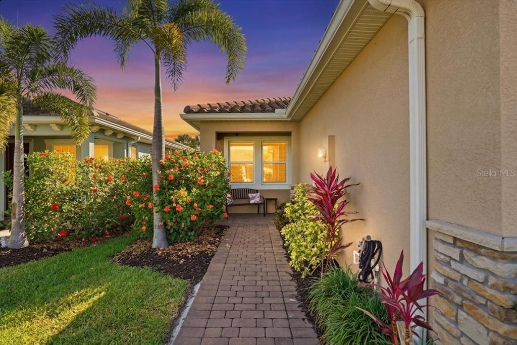 A brick walkway leads to a house entrance with a bench and tropical landscaping — 20406 GALILEO PLACE, VENICE, FL