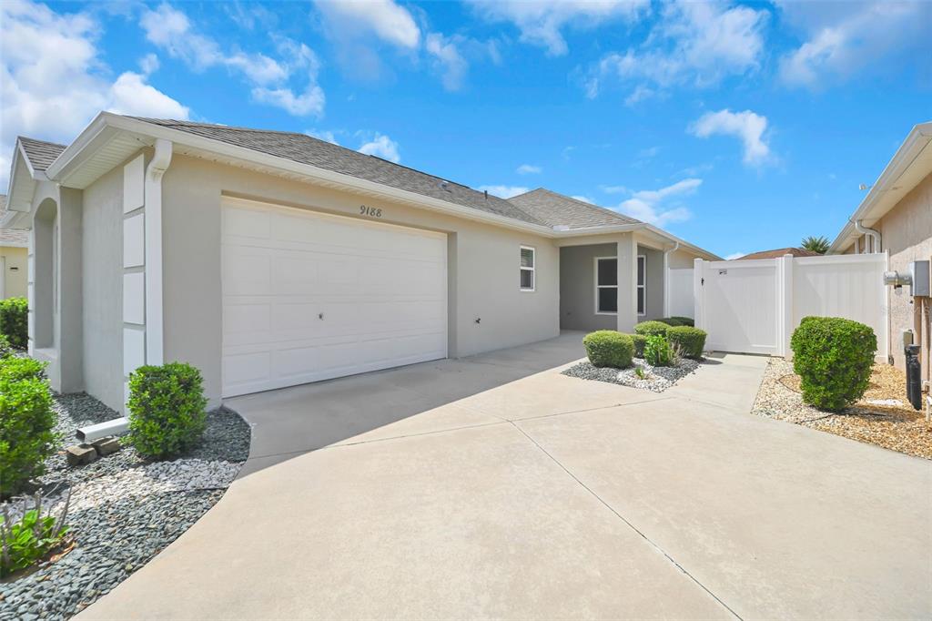 A beige house exterior features a two-car garage, a concrete driveway, and a white fence — 9188 SE 166TH SPRUNG LANE, THE VILLAGES, FL