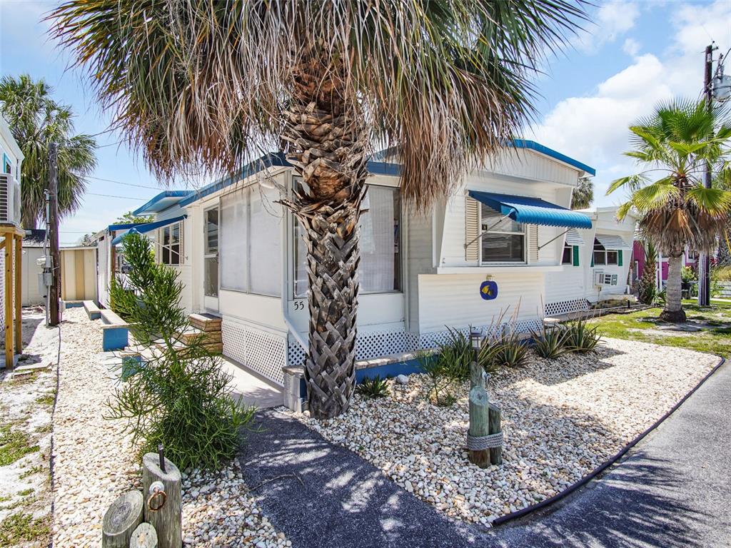 A white mobile home with blue awnings stands behind a palm tree and gravel yard — 800 CHESAPEAKE DRIVE UNIT 55, TARPON SPRINGS, FL