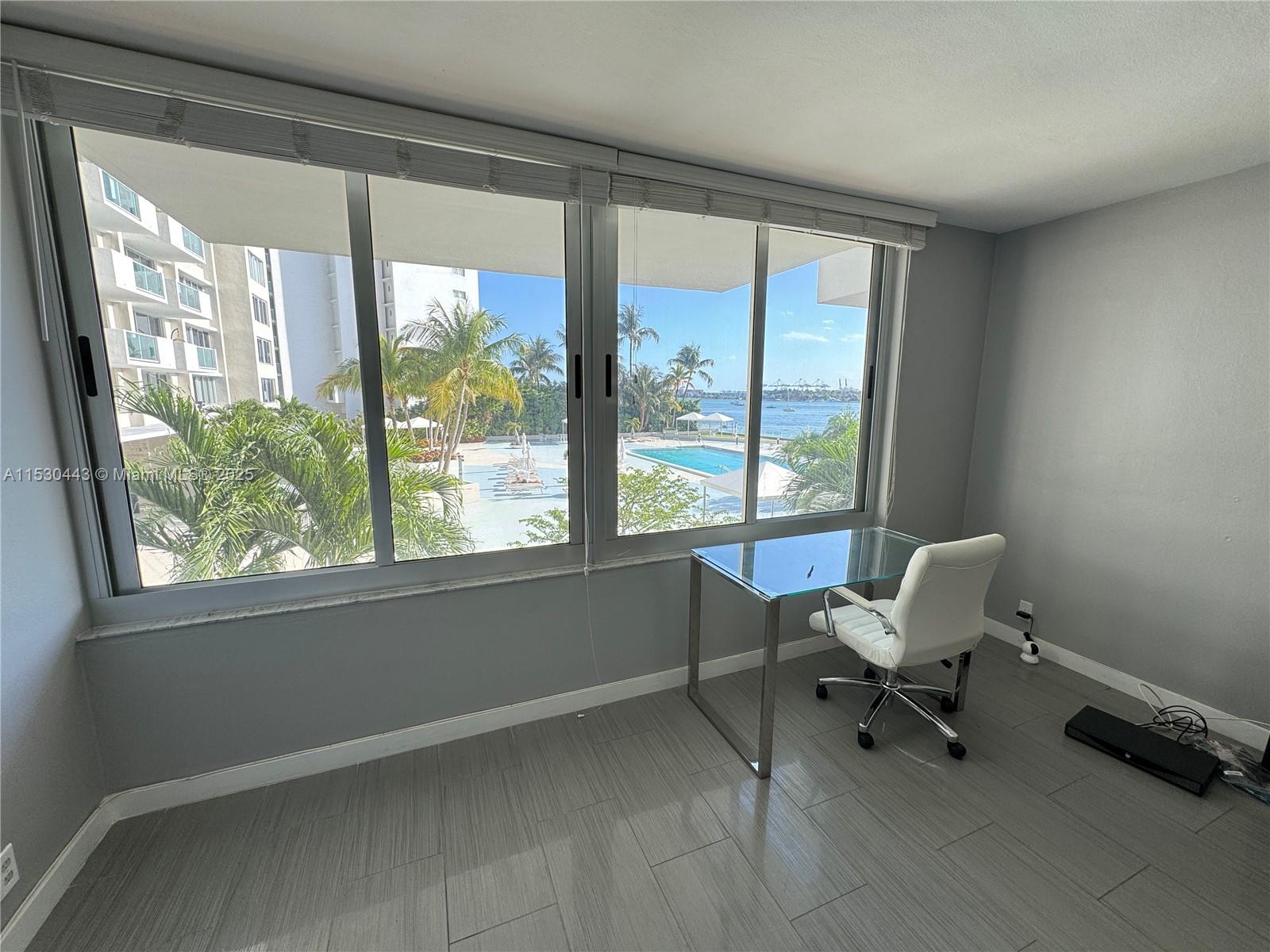 A home office features a glass desk, white chair, gray tile floors, and a waterfront view — 1200 West Ave # 202, Miami Beach FL 33139, MIAMI BEACH, FL