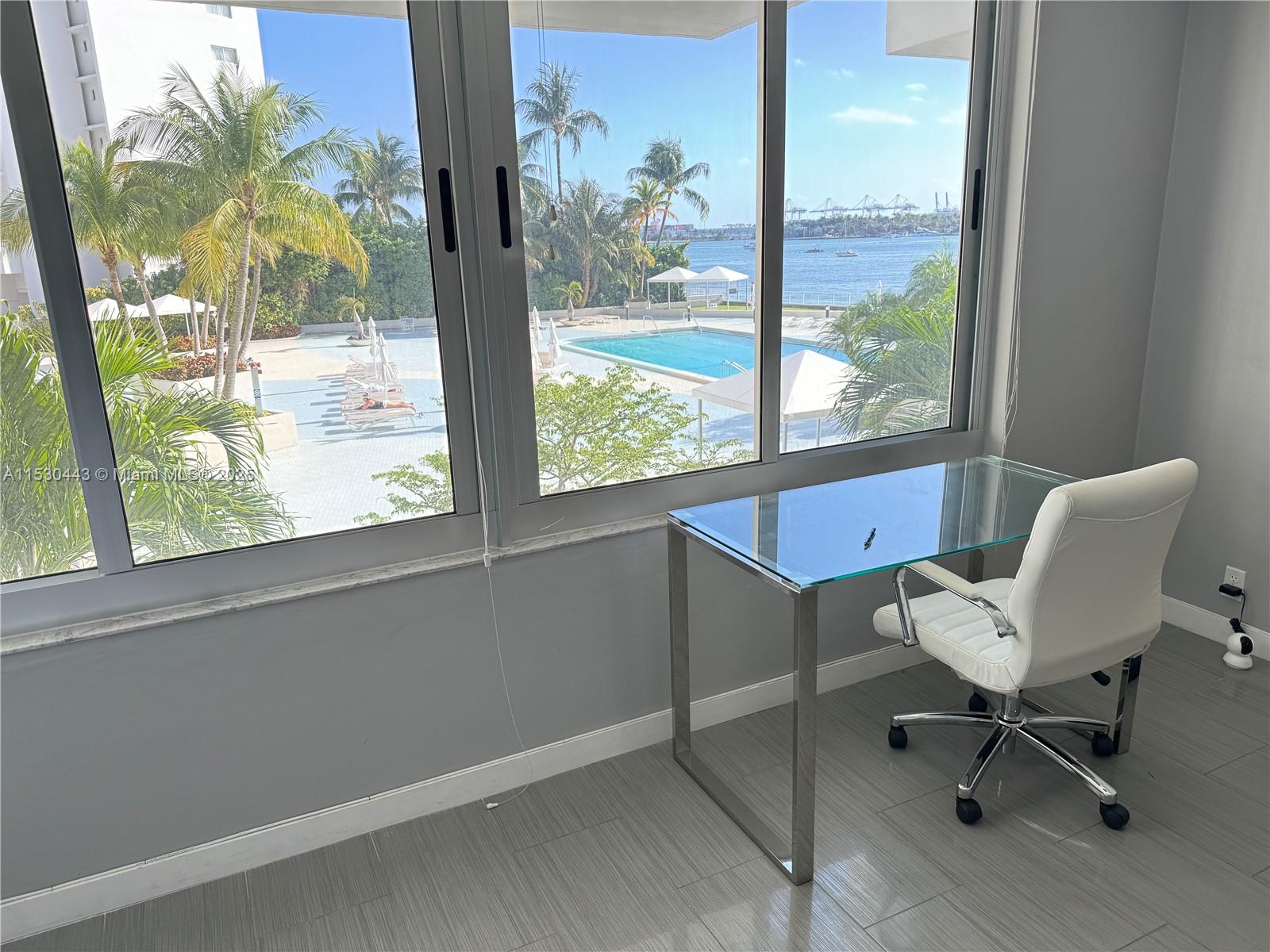 A desk and chair sit before a large window overlooking a swimming pool and bay — 1200 West Ave # 202, Miami Beach FL 33139, MIAMI BEACH, FL