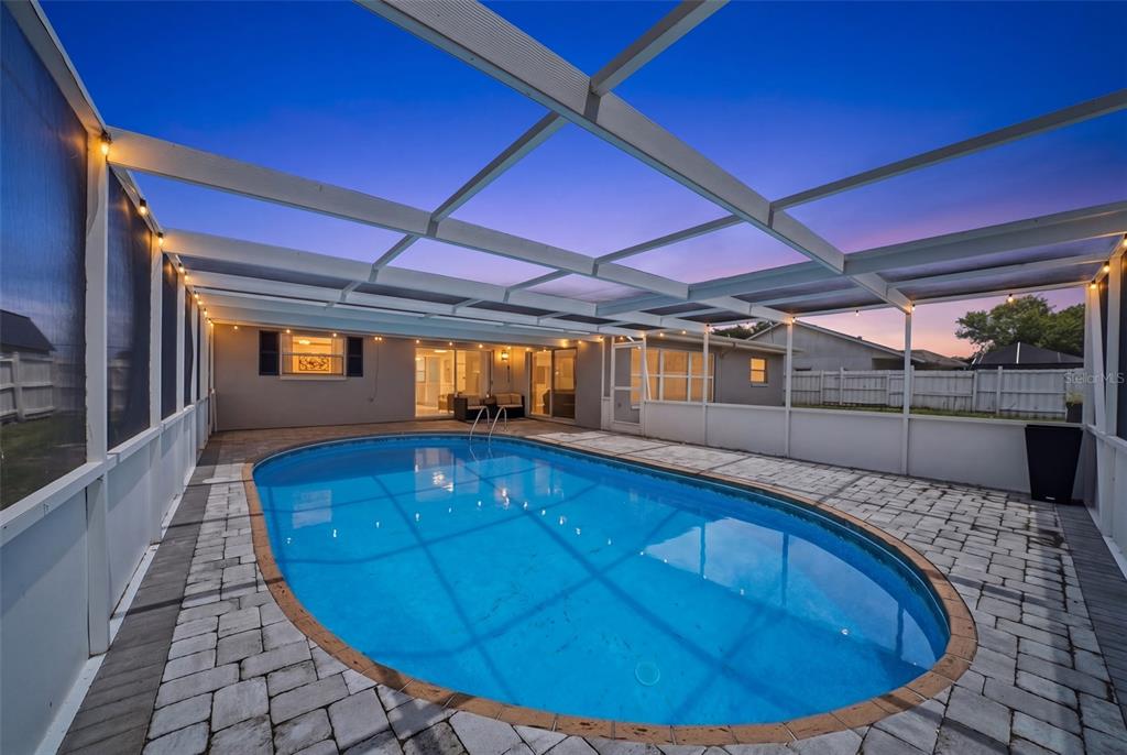 A backyard swimming pool with a screened-in enclosure and paver patio at dusk — 6072 AIRMONT DRIVE, SPRING HILL, FL