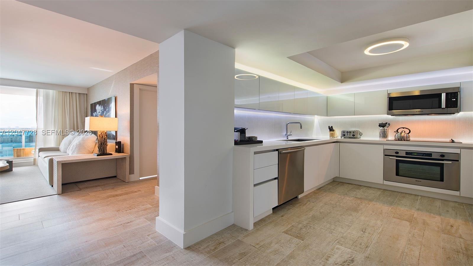 A modern kitchen with white cabinetry and stainless steel appliances opens into a living area — 102 24th St # 1245, Miami Beach FL 33139, MIAMI BEACH, FL