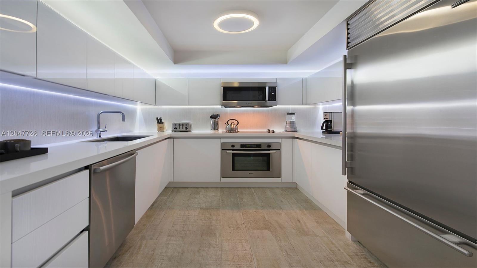 A modern, U-shaped kitchen features white cabinetry, stainless steel appliances, and wood-look tile flooring — 102 24th St # 1245, Miami Beach FL 33139, MIAMI BEACH, FL