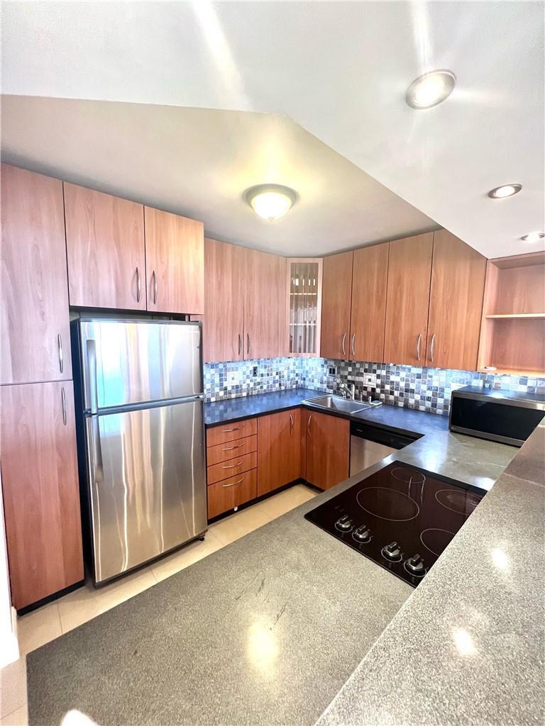 A kitchen features light wood cabinets, stainless steel appliances, dark countertops, and a tiled backsplash — 600 Three Islands Boulevard # 1108, Hallandale Beach FL 33009, HALLANDALE BEACH, FL