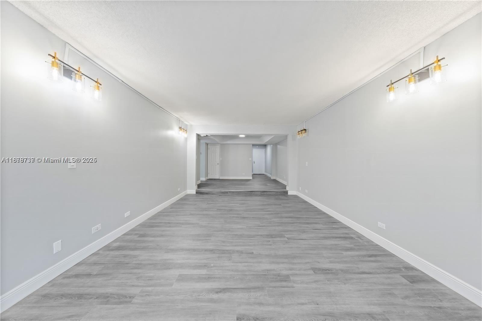 A long, empty room features gray wood-look flooring, light gray walls, and gold wall sconces — 3680 Inverrary Dr # 3J, Lauderhill FL 33319, LAUDERHILL, FL