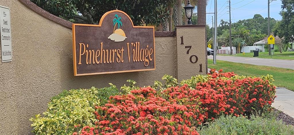 A sign for Pinehurst Village stands in front of a garden with red flowers — 1701 PINEHURST ROAD UNIT 16D, DUNEDIN, FL