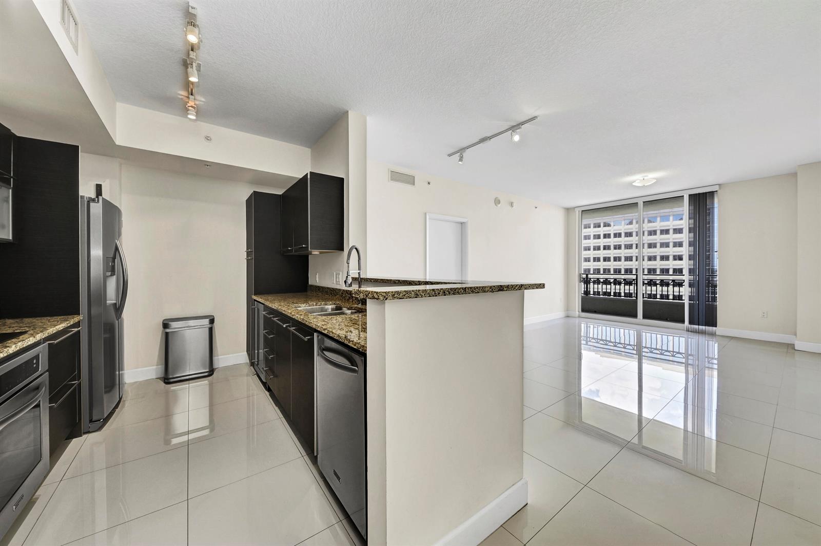 A modern kitchen with dark cabinets and granite countertops opens into a bright living area — 550 Okeechobee Boulevard # 1404, West Palm Beach FL 33401, WEST PALM BEACH, FL