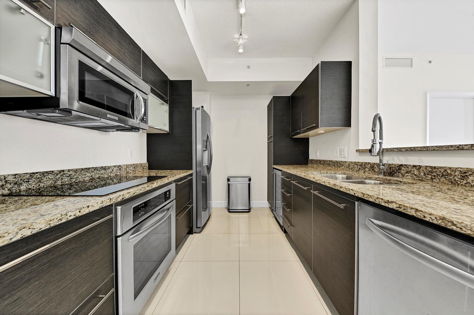A galley kitchen features dark wood cabinets, granite countertops, stainless steel appliances, and tile flooring — 550 Okeechobee Boulevard # 1404, West Palm Beach FL 33401, WEST PALM BEACH, FL