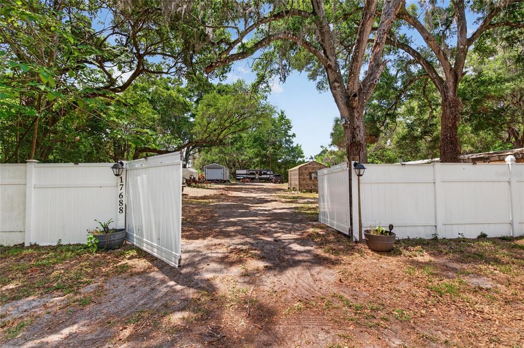 A dirt driveway leads through an open white vinyl gate toward a shed and trailer — 17684 NE 24TH AVENUE, CITRA, FL