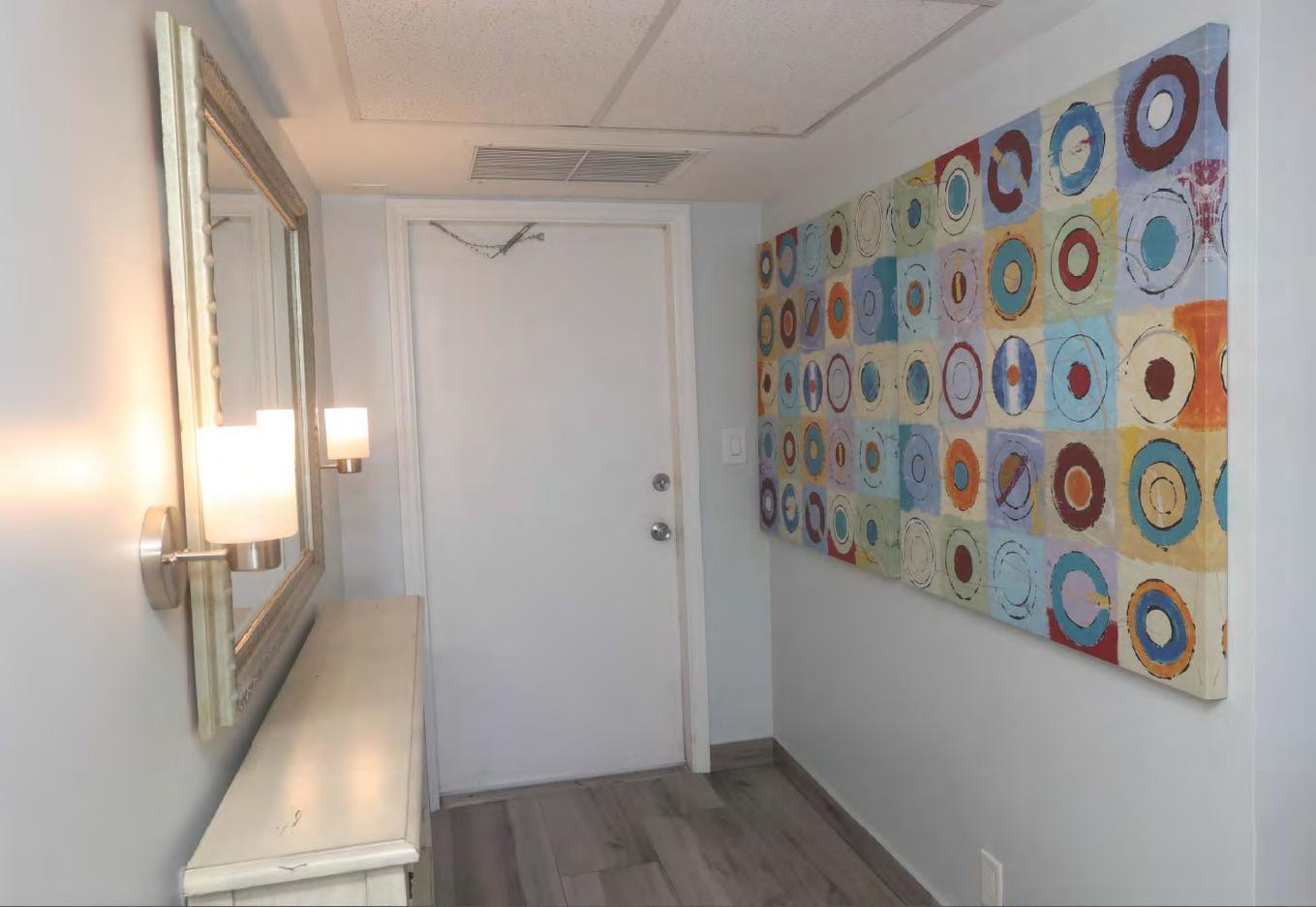 A hallway features a white door, a mirror above a console table, and colorful abstract art — 3091 N Course Drive # 303, Pompano Beach FL 33069, POMPANO BEACH, FL