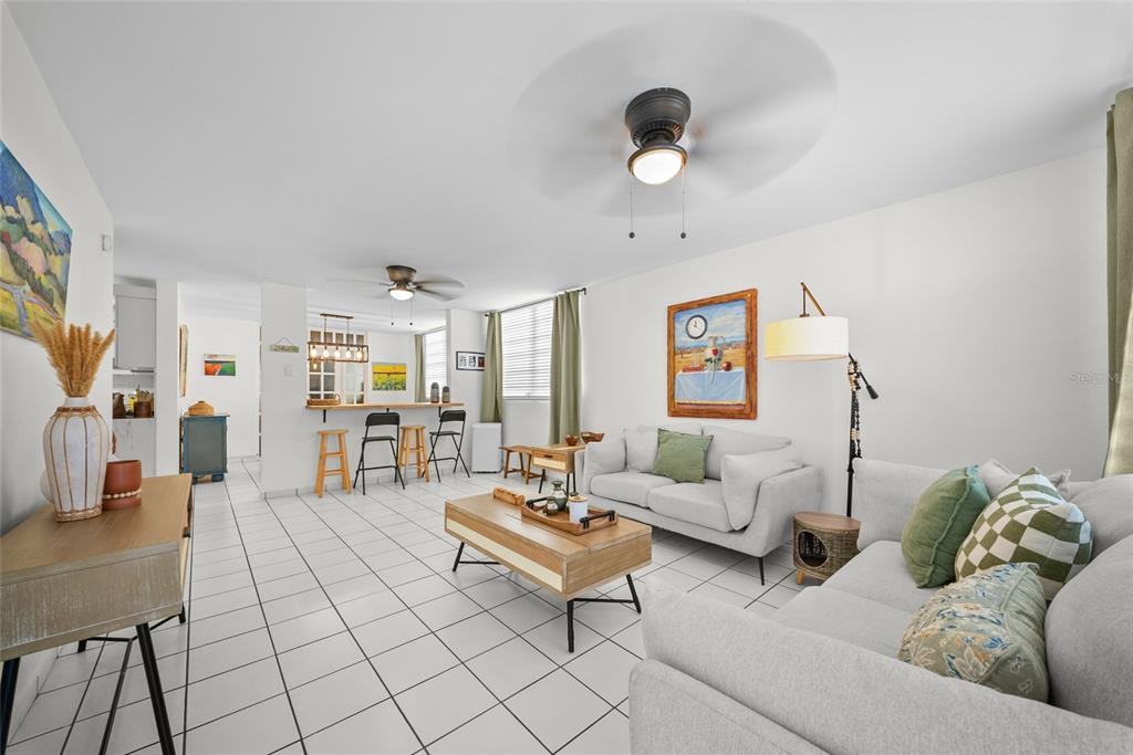 A living room with white tile floors, two grey sofas, and an open kitchen area — N13 CALLE BORIKEN URB. ALTURAS DEL ENCANTO, JUANA DIAZ, PR