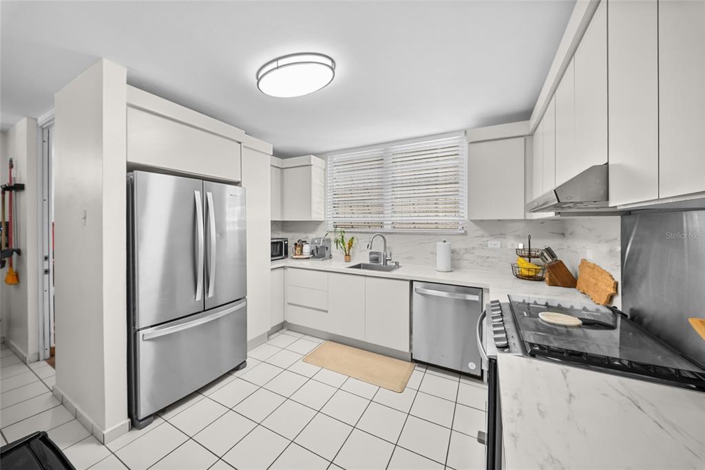 A modern kitchen features white cabinets, stainless steel appliances, and white tiled flooring — N13 CALLE BORIKEN URB. ALTURAS DEL ENCANTO, JUANA DIAZ, PR