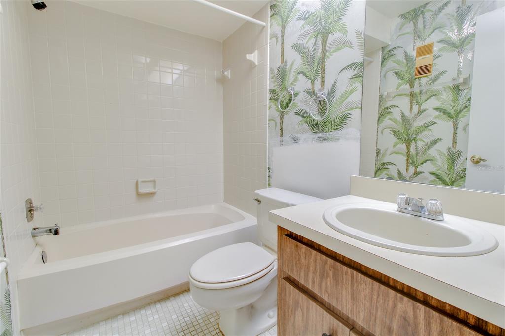 A bathroom features a white tub, a toilet, a wood vanity, and palm-patterned wallpaper — 3293 WESTRIDGE BOULEVARD UNIT 203, ORLANDO, FL