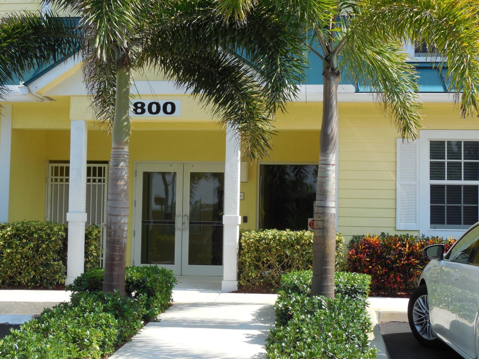 A yellow building exterior features a central entrance with double glass doors and palm trees — 800 Juno Ocean Walk # 301a, Juno Beach FL 33408, JUNO BEACH, FL