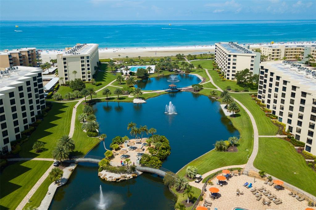 An aerial view of a beachfront resort complex featuring multiple buildings surrounding a central pond — 5780 MIDNIGHT PASS ROAD UNIT 210, SARASOTA, FL