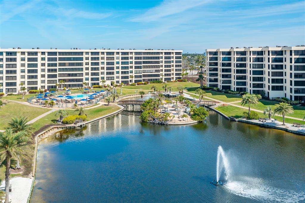 An aerial view shows two large apartment buildings surrounding a lake with a central fountain — 5780 MIDNIGHT PASS ROAD UNIT 210, SARASOTA, FL